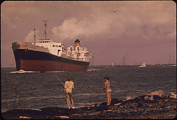 Couple fishing in a polluted river while a oil freighter sails by in the background.