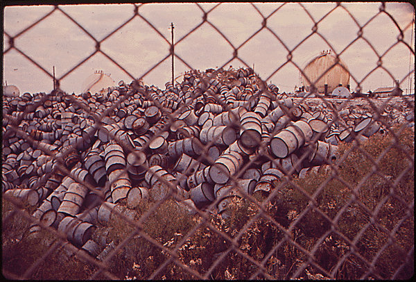 A large pile of used oil drums lies in the open air near an Exxon refinery
