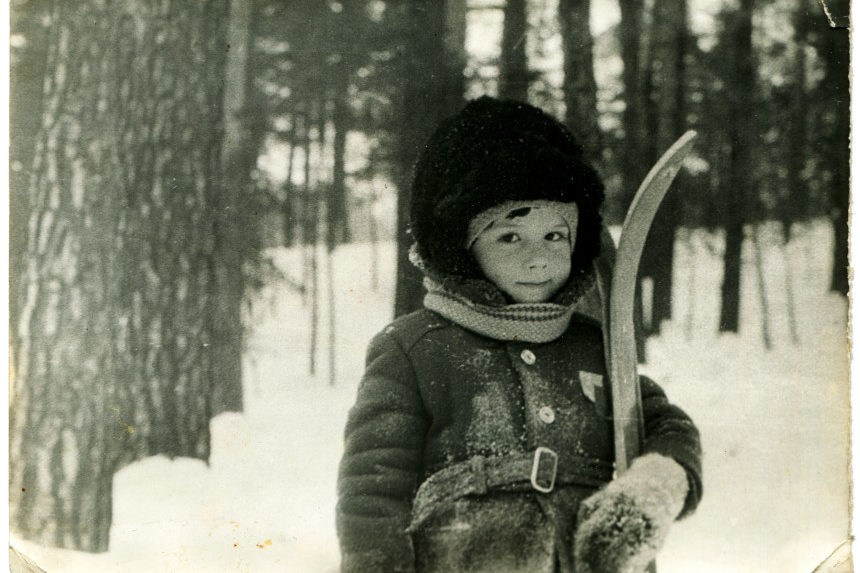 A boy with a ski stands in a snow covered forest in winter.
