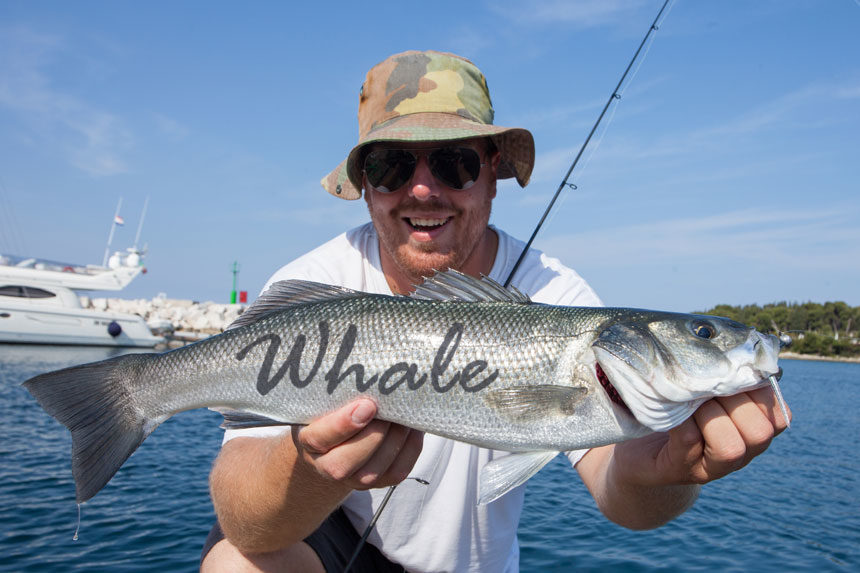 A man with a fishing rod holding a foot-long fish with the word WHALE written on it.