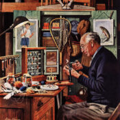 A gray-haired man in a blue cardigan sits at a desk, surrounded by the accouterment of fishing, preparing a fly with a pair of scissors.