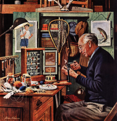 A gray-haired man in a blue cardigan sits at a desk, surrounded by the accouterment of fishing, preparing a fly with a pair of scissors.