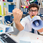An angry man at a messy office desk shakes his fist while shouting into a bullhorn