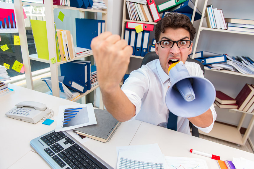 An angry man at a messy office desk shakes his fist while shouting into a bullhorn